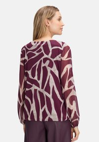 Kobieta z długimi blond włosami, ubrana w przezroczystą bluzkę z długim rękawem w abstrakcyjny fioletowy i beżowy wzór, stojąca bokiem na tle jednolitym.