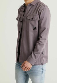 Homme portant une veste mauve boutonnée avec des poches poitrine et un jean bleu clair, debout devant un fond clair et uni.