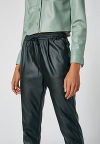 Chemise cropped en simili cuir vert avec un col rabattu et des boutons pression, associée à un pantalon de jogging noir à taille élastique avec texture.