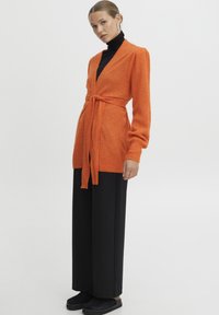 Oranžinė megzta kardiganas su ilgomis rankovėmis ir juosmens raiščiu, dėvimas virš juodo polo megztinio, derinamas su juodomis plačiomis kelnių ir tamsiais bateliais.