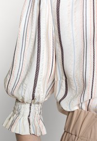 Blusa riscada em tecido leve, apresentando mangas bufantes, punhos franzidos e cores pastel alternadas com riscas escuras.