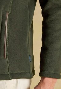 Primo piano di una persona che indossa una giacca in pile verde oliva, con una tasca laterale visibile e un'etichetta del marchio Joules cucita, pantaloni beige mostrati parzialmente.