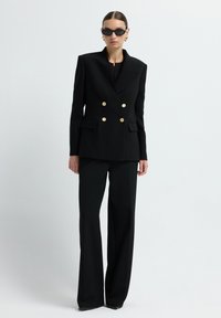 Vrouw in een zwarte dubbelzijdige blazer met gouden knopen, bijpassende wijde broek, zwarte zonnebril en gouden creolen tegen een witte achtergrond.