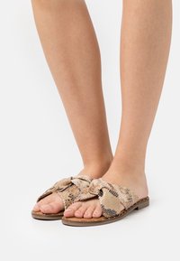 Płaskie sandały na krokodyl, wykonane z fakturowanego materiału w wężowy wzór w odcieniach beżu i brązu, z dużym dekoracyjnym węzłem z przodu.