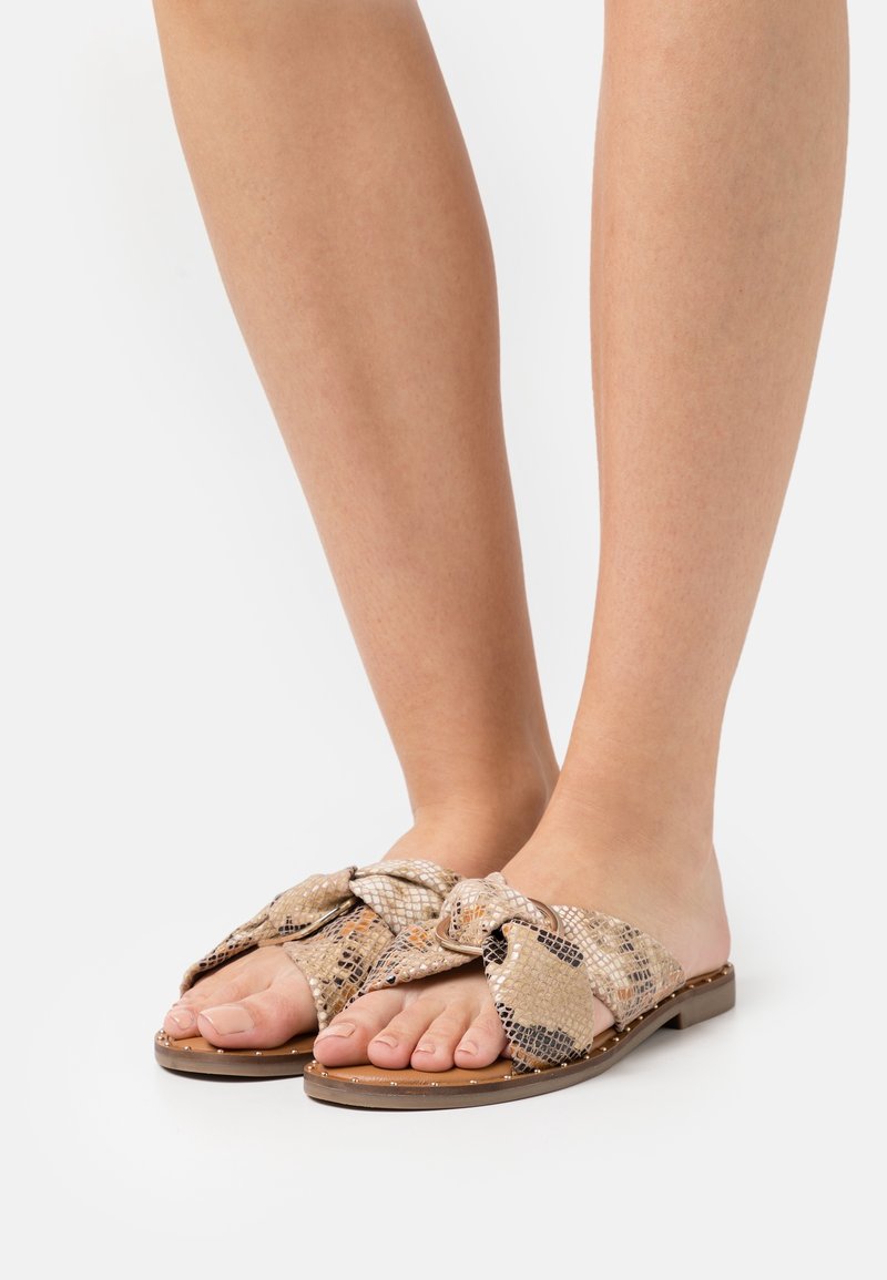 Płaskie sandały na krokodyl, wykonane z fakturowanego materiału w wężowy wzór w odcieniach beżu i brązu, z dużym dekoracyjnym węzłem z przodu.