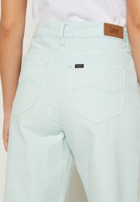 Jeans de ganga de um azul claro com uma cintura alta, bolsos traseiros e um rótulo em couro castanho na cintura. Textura suave e ajuste casual.