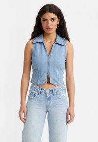 Colete cropped sem mangas em denim azul claro com fecho frontal, gola pontuda e bainha arredondada. Combinado com jeans claros desgastados.