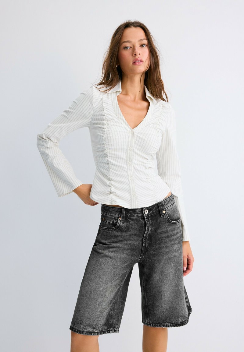 Femme aux longs cheveux porte un chemisier blanc froncé boutonné et un short en jean noir délavé longueur genoux sur un fond clair uni.