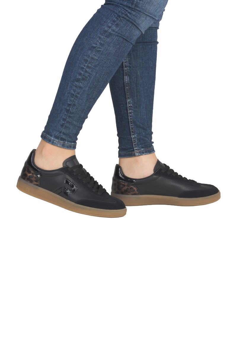 Personne portant un jean skinny bleu et des baskets noires avec des accents en imprimé léopard marron et des semelles en gomme, debout sur un fond blanc.