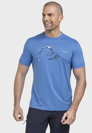 Lächelnder Mann mit kurzen Haaren, der ein hellblaues kurzärmeliges Hemd mit einem Bergmotiv trägt, kombiniert mit dunkler Hose.