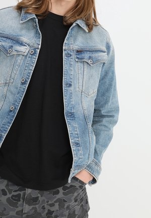 Persona vistiendo una chaqueta de mezclilla azul claro sobre una camisa negra y pantalones de camuflaje grises, de pie con una mano en el bolsillo contra un fondo blanco.