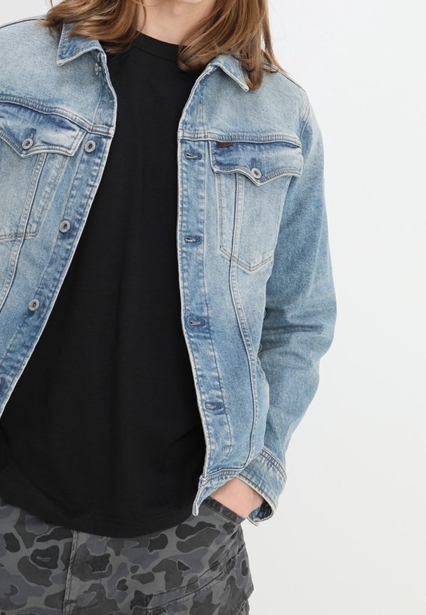 Person med ljusblå denimjacka över svart skjorta och grå kamouflagedbyxor, står med en hand i fickan mot vit bakgrund.