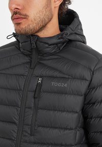 Black puffer jacket with a hood, featuring quilted texture, front zipper, and a chest pocket. Logo printed on the left side.