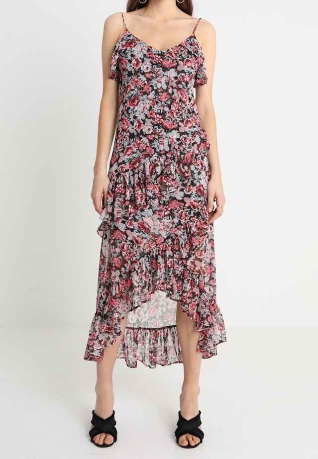 Femme portant une robe sans manches à imprimé floral avec des volants et un ourlet asymétrique, associée à des sandales noires à talons.