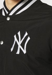 Black varsity jacket with a ribbed black and white collar, featuring a prominent white embroidered logo on the left side. Metal snap closures.
