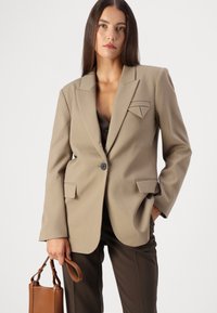 Veste beige ajustée avec col châle, deux poches avant et un détail en triangle sur le revers gauche. Associée avec un pantalon marron et un sac à main.