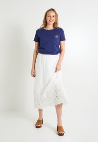 Camiseta de color azul marino con mangas cortas y botones en los hombros, combinada con una falda blanca de bordado con ojales que presenta un dobladillo ondulado.