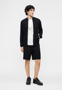 Mladi moški z vinskimi kodri, oblečen v črni pulover s patentnim zadrgo, belo majico z potiskom, črne kratke hlače in črne superge, stoji z eno roko v žepu.