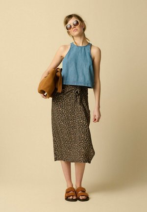 Femme portant des lunettes de soleil rondes, un top en denim sans manches, une jupe midi à imprimé léopard, des sandales marron cloutées, tenant un sac marron sur fond beige.