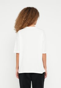 Λευκό βαμβακερό t-shirt με άνετη εφαρμογή, κοντά μανίκια και στρογγυλό λαιμό. Η πίσω όψη εμφανίζει ελάχιστες ραφές. Συνδυασμένο με μαύρο παντελόνι.