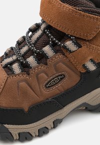Braune Wildleder-Wanderstiefel mit dunkler Gummisohle, Mesh-Akzenten und kontrastierendem Schnürsystem. Verfügt über ein Logo an der Seite und strukturierte Oberflächendetails.