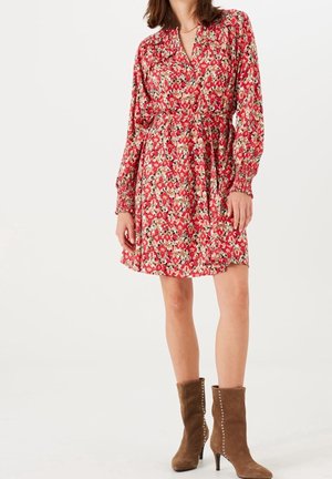 Femme portant une robe rouge à fleurs à manches longues avec une ceinture, assortie à des bottes en daim marron à talons mi-mollet sur fond blanc.