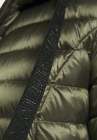 Zwarte gestructureerde band met reliëftekst over een olijfgroene glanzende gewatteerde stof, waarschijnlijk onderdeel van een gewatteerde jas of tas.