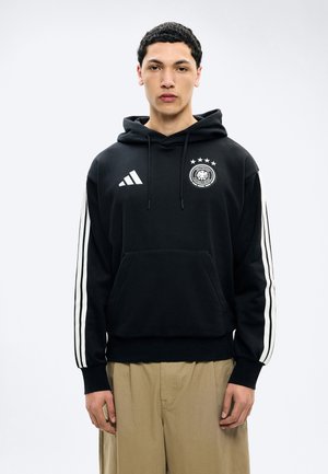 Jaunas vyras dėvi juodą Adidas gobtuvą su baltomis juostelėmis ir Vokietijos futbolo asociacijos logotipu, stovintis priešais šviesų, vienodu foną.