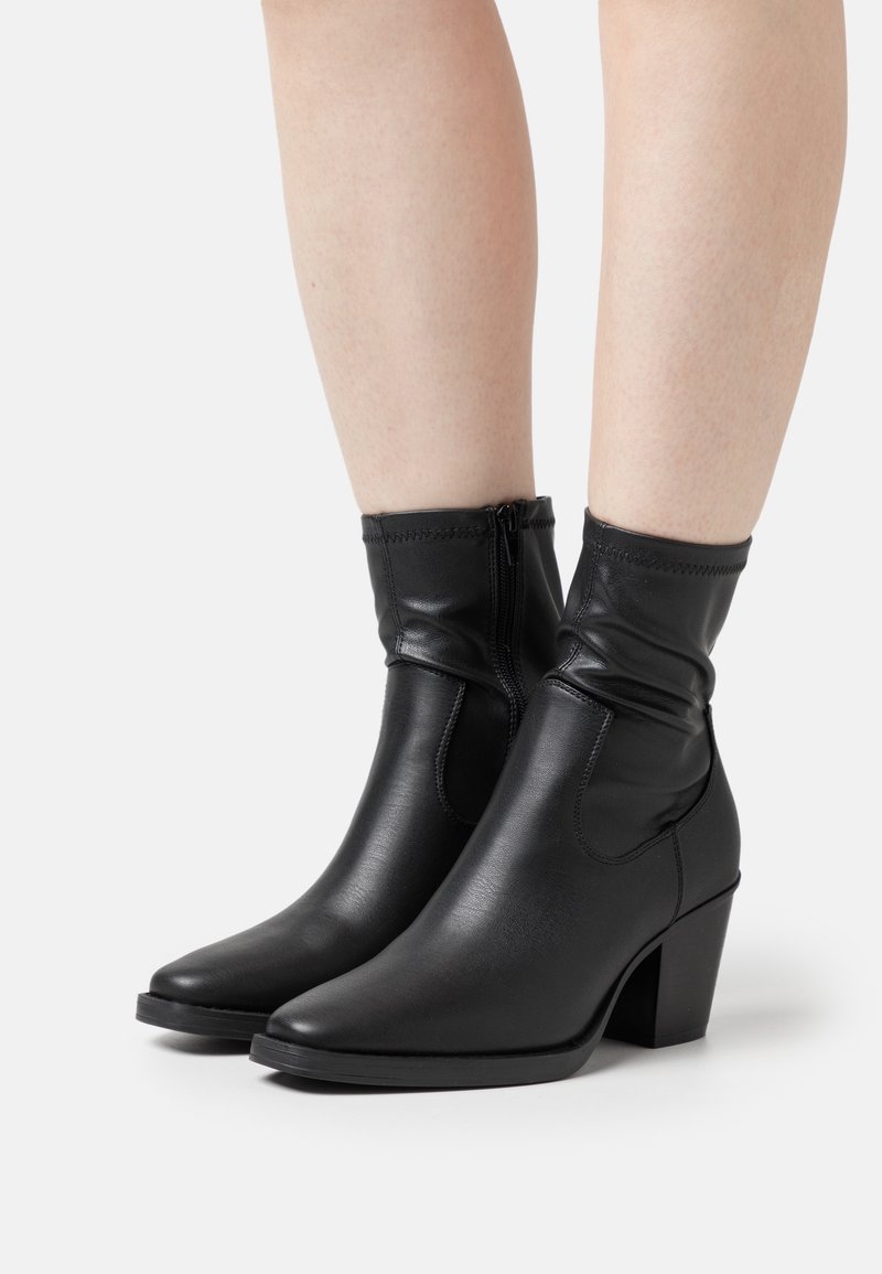 Bottes en cuir noires mi-mollet avec fermeture éclair latérale et talons carrés portées sur jambes nues sur fond blanc.