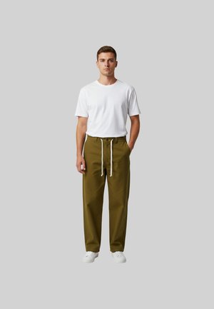 Giovane uomo in piedi, dritto, con una maglietta bianca, pantaloni verde oliva con coulisse e scarpe da ginnastica bianche, su uno sfondo grigio uniforme.