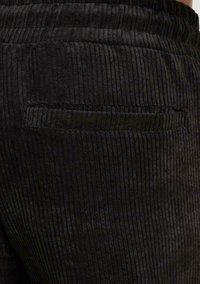 Sort cordstof, der viser lodrette ribber, med en detalje i baglommen. Teksturen virker blød og fyldig, hvilket fremhæver komfort.