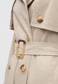 Trench-coat beige avec une taille cintrée par une ceinture, de gros boutons ronds au design dégradé, et un col structuré. Texture de tissu lisse et durable.