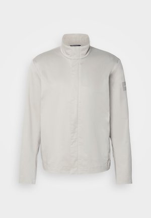 Veste longue à manches longues gris clair avec col haut, texture lisse, fermeture avant et logo discret sur la manche. Design minimaliste.