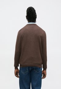 Barna kötött kardigán bordázott mandzsettával és szegéllyel, egyszerű dizájnnal és kerek nyakkivágással, kék farmernadrággal párosítva.