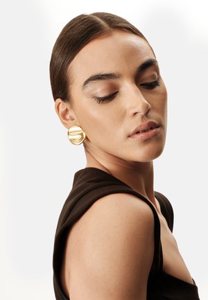 Pendientes circulares de oro con acabado pulido, con un diseño ondulado. La modelo lleva una blusa sin mangas de color marrón oscuro.