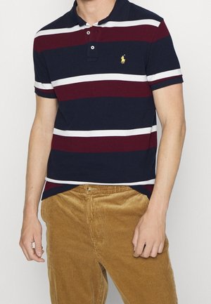 Polo-shirt rayé bleu marine et bordeaux avec bandes blanches, trois boutons au col, petit logo jaune. Porté avec un pantalon en velours côtelé beige.