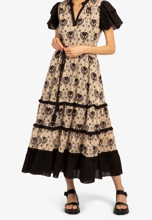 Frau in einem beige-schwarzen gemusterten Midikleid mit Puffärmeln und schwarzen Sandalen, steht vor einem einfarbigen Hintergrund.
