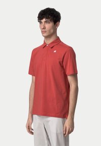 Polo rouge à manches courtes, doté d'un col à boutons et d'un logo blanc sur le côté gauche de la poitrine. Confectionné en tissu léger.