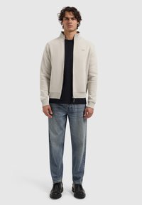 Light grey zip-up jacket over a black shirt, paired with loose-fitting blue jeans and black shoes. Features a high collar and ribbed cuffs.