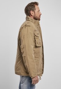 Jachetă de stil militar bej, cu multiple buzunare, închidere cu fermoar și nasturi, epauleți pe umeri și o croială lejeră. Fabricată dintr-un material durabil.