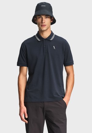 Giovane uomo che indossa una polo blu navy con righe bianche sul colletto e un cappello bucket nero, in piedi con una mano in tasca contro uno sfondo semplice.