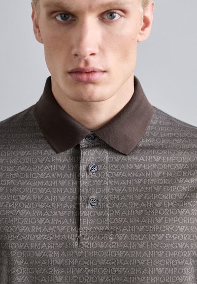 Brūns polo krekls ar strukturētu rakstu, kurā ir "Emporio Armani" zīmols. Tam ir apkakles kakla izgriezums un trīs tumši pogas.