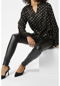 Femme assise portant un chemisier noir à motif de feuilles dorées, des leggings en cuir noirs et des chaussures à talons ouvertes arrière noires à bouts pointus.