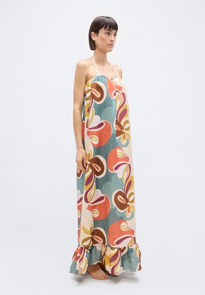 Γυναίκα όρθια, ξυπόλητη, φορώντας ένα πολύχρωμο, φλοράλ μακρύ φόρεμα χωρίς τιράντες με βολάν στο τελείωμα, μπροστά σε ένα απλό φωτεινό φόντο.