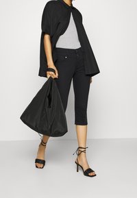 Persona vestida con pantalones cortos negros, camiseta gris, camisa negra abierta, sandalias de tacón negro con tiras, sosteniendo un gran bolso negro de mano.