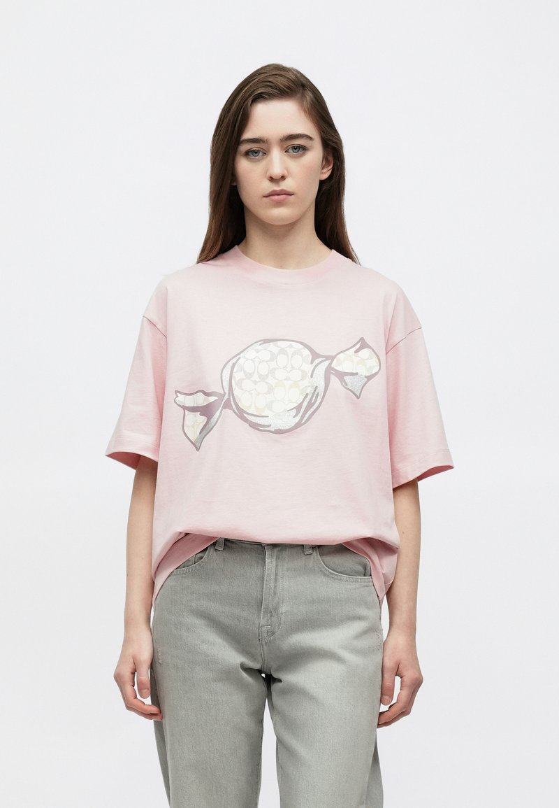 Ροζ βαμβακερό μπλουζάκι με γραφικό σχέδιο καραμέλας σε λευκό και γκρι. Χαλαρή εφαρμογή, κοντά μανίκια, στρογγυλή λαιμόκοψη. Φορεμένο με γκρι τζιν παντελόνι.