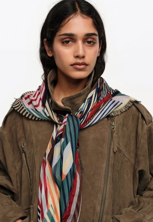 Mujer joven de cabello oscuro que lleva una chaqueta de ante marrón y una bufanda a rayas multicolor atada alrededor del cuello, mirando hacia adelante.
