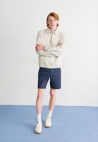 Lys beige bomberjakke med elastisk talje, marineblå shorts, hvide sneakers og ankelsokker. Glat stof, afslappet pasform, minimalistisk design.