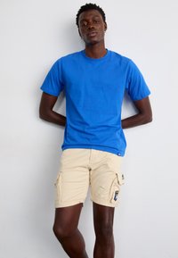 Tricou din bumbac albastru cu guler rotund, asociat cu pantaloni scurți cargo bej cu buzunare și margine rulat. Material neted și croială confortabilă.