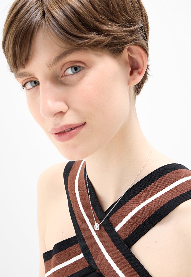 Collier en argent avec un pendentif carré, porté par un mannequin en haut rayé marron et noir avec des accents blancs, en mettant l'accent sur le décolleté.